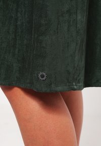Falda de terciopelo verde oscuro con una textura suave, que presenta un pequeño detalle de logo gris cerca del dobladillo. Acabado suave y diseño casual.