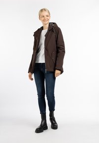 Brun vandtæt jakke med hætte, der har lynlås foran, sidelommer og en let oversized pasform, parret med mørke jeans og sorte ankelstøvler.