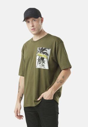 T-shirt en coton vert olive avec un motif graphique de palmier sur la poche, accompagné de texte jaune. Casquette noire avec le nom de la marque embossé à l'avant.