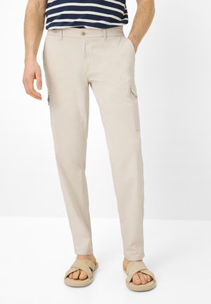 Mann trägt beige Cargo-Hosen mit Seitentaschen, ein marineblau-weiß gestreiftes Hemd und beige, gekreuzte, offene Sandalen, steht mit einer Hand in der Tasche.