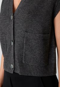 Dunkelgraue ärmellose Strickjacke mit Taschen und Knöpfen, getragen über schwarzen Hosen.