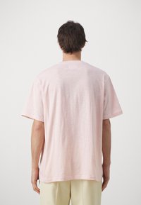 Camiseta de manga corta en rosa claro, hecha de tela ligera, con cuello redondo y un ajuste relajado. Textura suave con un diseño mínimamente detallado.