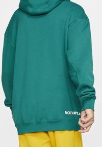 Vihreä ylipitkä huppari, jossa on pehmeä pinta, huppu ja suuri etupuolella oleva kengurutasku. Alareunassa on valkoinen "EI OLE NÄYTE" -teksti.