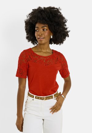 Mujer con cabello rizado que lleva una blusa de encaje roja, pantalones blancos y cinturón marrón, con la mano en el bolsillo, mirando hacia un lado.