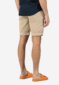 Beige Baumwollshorts mit umgeschlagenen Säumen, Rückentasche, getragen mit orangefarbenen Sandalen. Modell von hinten mit einem marineblauen Shirt abgebildet.