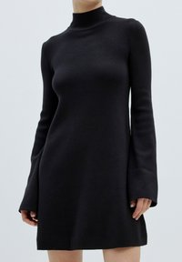 Zwarte jurk met lange mouwen en een hoge hals, gemaakt van een zachte stof met uitlopende mouwen. De jurk heeft een slim fit en een uitlopende onderrand.