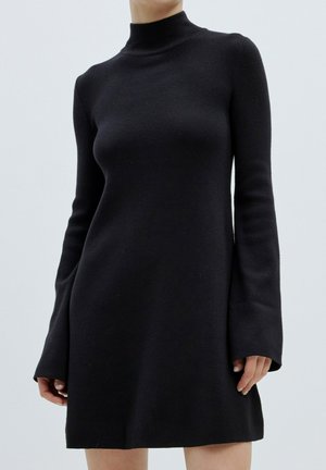 Zwarte jurk met lange mouwen en een hoge hals, gemaakt van een zachte stof met uitlopende mouwen. De jurk heeft een slim fit en een uitlopende onderrand.