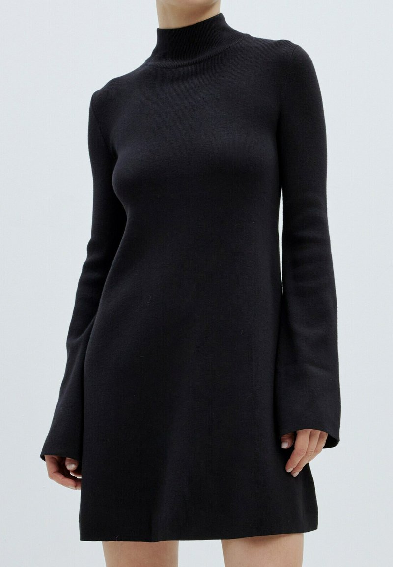 Zwarte jurk met lange mouwen en een hoge hals, gemaakt van een zachte stof met uitlopende mouwen. De jurk heeft een slim fit en een uitlopende onderrand.