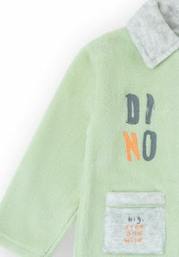 Chaqueta de forro polar verde claro con cuello gris, con texto "DINO" en gris oscuro y naranja, y un bolsillo suave gris con la inscripción "grande, libre y salvaje".