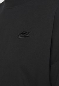 Svart bomullströja med hög hals som har en texturerad yta och en liten, präglad svart Nike-logga på övre vänstra sida.