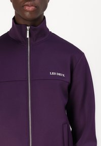 Chaqueta morada con cremallera, hecha de material sintético, con cuello alto, bolsillos laterales y un logo blanco en el pecho. Textura suave.