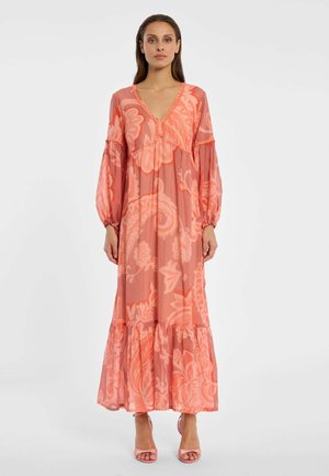 Frau trägt ein langes Pfirsichkleid mit Blumenmuster, langen Puffärmeln, V-Ausschnitt und gerafftem Saum, kombiniert mit pfirsichfarbenen Sandaletten mit hohen Absätzen.