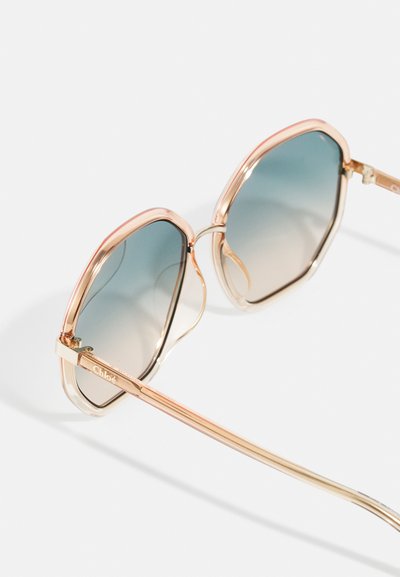 Occhiali da sole con montatura in oro rosa e grandi lenti esagonali blu sfumate poggiati su una superficie bianca.