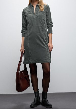 Kobieta w szarej sukience z długim rękawem z kieszeniami na piersi, czarnych rajstopach, czarnych butach i trzymająca brązową skórzaną torebkę przy boku.