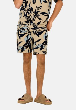 Hombre con pantalones cortos beige con estampado floral y camisa de manga corta a juego, con sandalias marrones abiertas, de pie sobre fondo blanco.