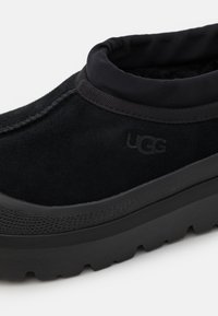 Black suede slip-on shoe with a padded collar, featuring a textured black rubber sole and the UGG logo embossed on the side.
