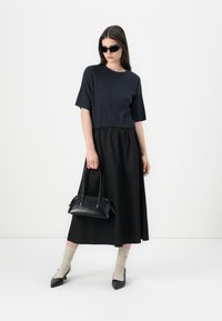 Svart ribbet topp med korte ermer, svart flytende midi-skjørt, svart håndveske, beige sokker og svarte spisse hæler. Minimalistisk design og farger.