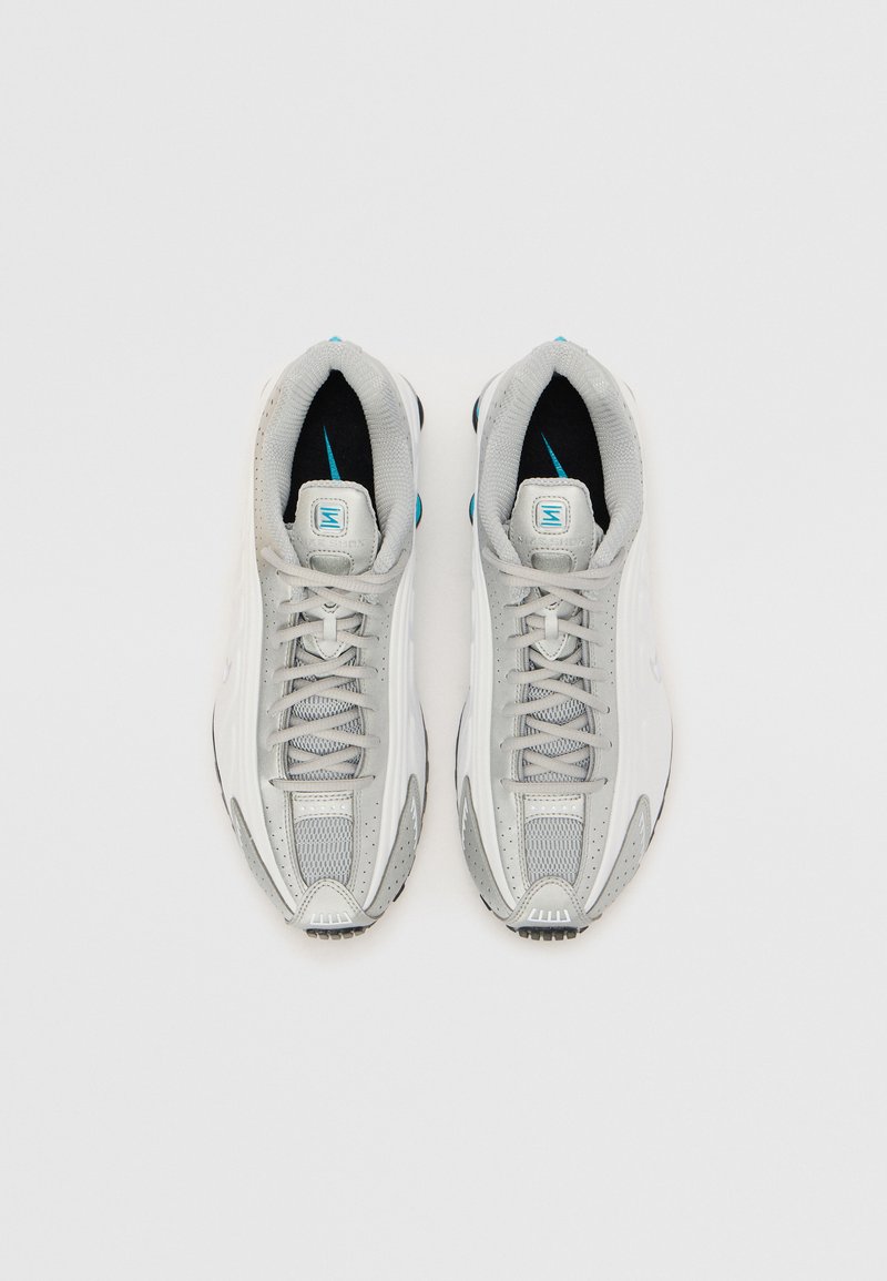 Zapatillas deportivas de color gris claro, fabricadas con materiales de malla y sintéticos, que presentan una superficie texturizada, un diseño con cordones y acentos en azul en la lengüeta.