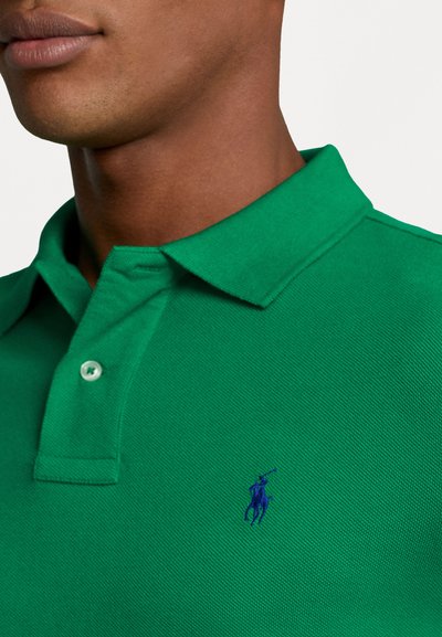 Zaļš polo krekls no teksturēta auduma ar izšūtu zilu logotipu kreisajā pusē, standarta apkakle un divas pogas.