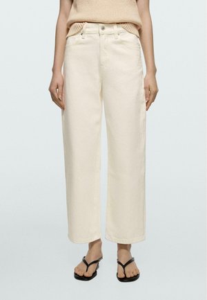 Femme portant un pantalon crème taille haute à jambes droites avec bouton et fermeture éclair à l'avant, associé à des sandales noires à lanière et un haut en maille beige.