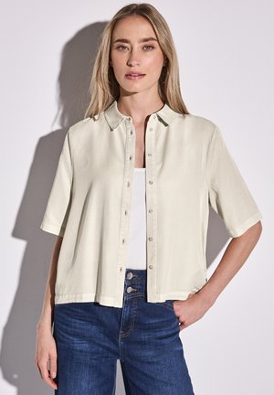 Lichtbeige overhemd met korte mouwen en knoopsluiting, gedragen over een witte tanktop. Gepaard met donkerblauwe jeans, met zichtbare knopen.