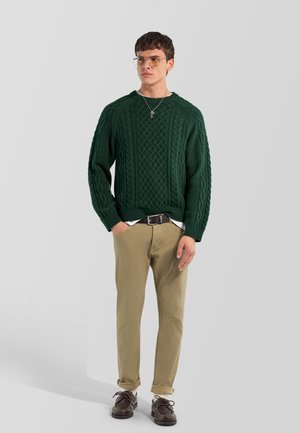 Joven con un jersey de punto grueso verde oscuro, pantalones beige con puños, zapatos marrones, gafas y un collar plateado, de pie con una mano en el bolsillo.