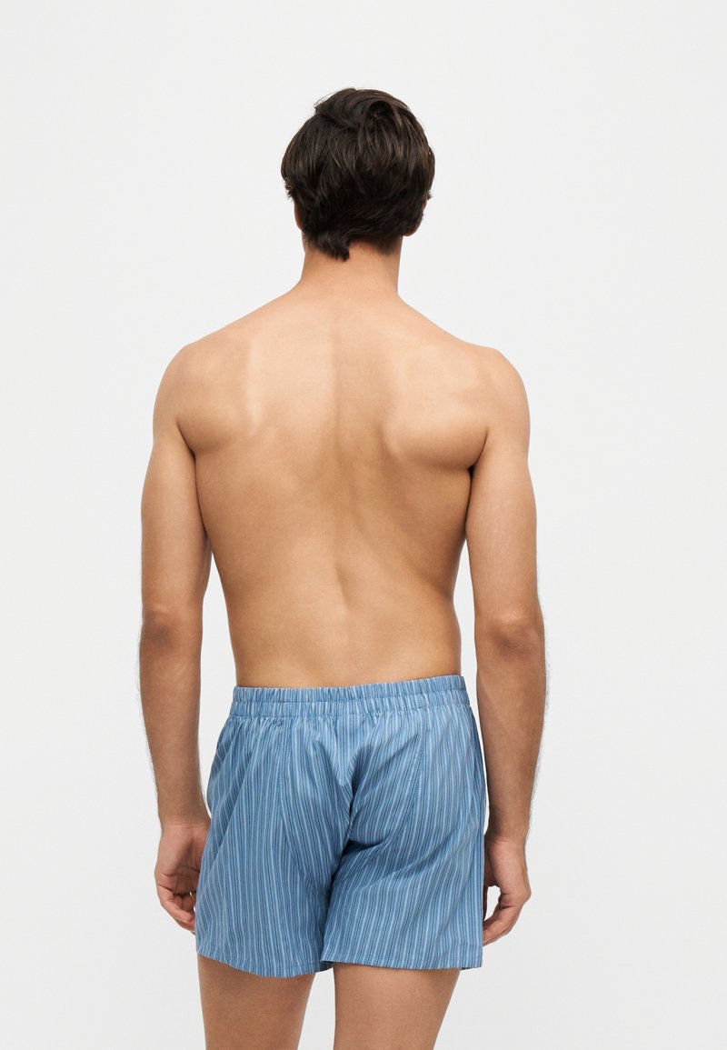 Mann steht mit dem Rücken zur Kamera, trägt blaue gestreifte Shorts mit elastischem Bund vor schlichtem hellem Hintergrund.