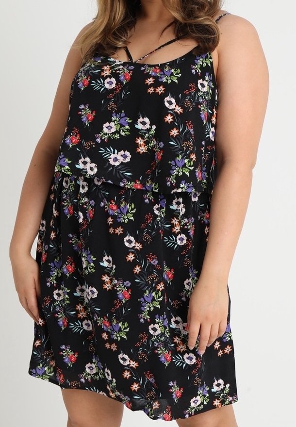 Femme portant une robe noire sans manches avec des motifs floraux violets, rouges, blancs et verts et des détails à volants superposés.