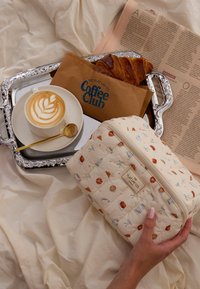 Najedwabna kremowa kosmetyczka z kolorowymi ilustracjami ciast i kawy, z górnym zamkiem błyskawicznym i metką marki z boku.