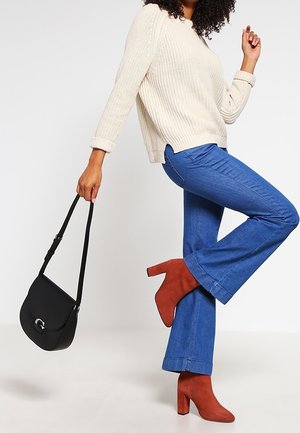 Beiger Strickpullover, blaue ausgestellte Jeans, rote knöchelhohe Stiefel mit Absatz und eine schwarze Schultertasche. Strukturiertes Gewebe und kräftige Farben.