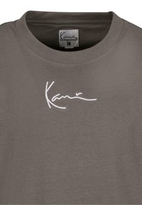 T-shirt en coton gris avec un col rond, présentant un logo brodé blanc sur la poitrine et une texture douce. Étiquette de taille : L.