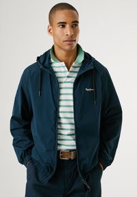 Chaqueta con capucha de color navy con puños elásticos, llevada sobre una camiseta tipo polo a rayas verdes y blancas. Presenta un logotipo en el pecho y un cinturón marrón.