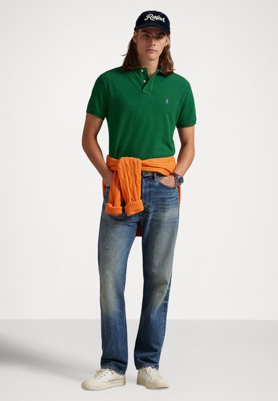 Πράσινο πόλο πουκάμισο, πορτοκαλί πουλόβερ δεμένο στη μέση, μπλε τζιν τζιν και κρεμ αθλητικά παπούτσια. Η καπέλο έχει λογότυπο σε λευκό χρώμα σε σκούρο φόντο.