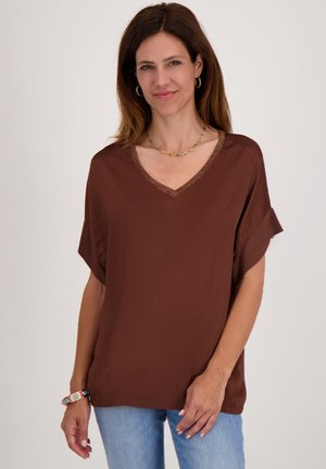 Bruine loszittende V-hals blouse met korte mouwen, voorzien van kantafwerking bij de hals. Gecombineerd met lichtblauwe jeans.