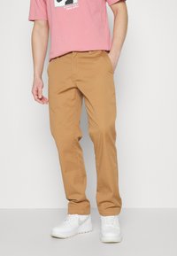 Persoon in tan rechte broek, een roze graphic t-shirt, en witte sneakers, staand tegen een effen lichte achtergrond.