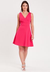 Vestido ajustado sin mangas en un vibrante color rosa, con escote en V, falda acampanada y una textura suave. Sandalias plateadas con tiras complementan el look.