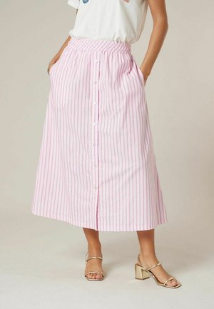 Femme portant une jupe midi à rayures roses et blanches, taille haute, boutonnée à l'avant avec des poches, et des sandales à talons blocs dorées avec brides.