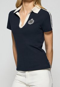 Kvinna iklädd en marinblå kortärmad polo med vit kant och ett emblem på bröstet, tillsammans med vita byxor mot en ljus bakgrund.