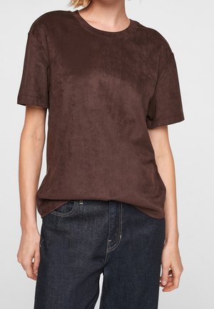 Person med en simpel brun kortærmet skjorte og mørkeblå jeans, hænder afslappet ved siden, mod en ensartet lys baggrund.