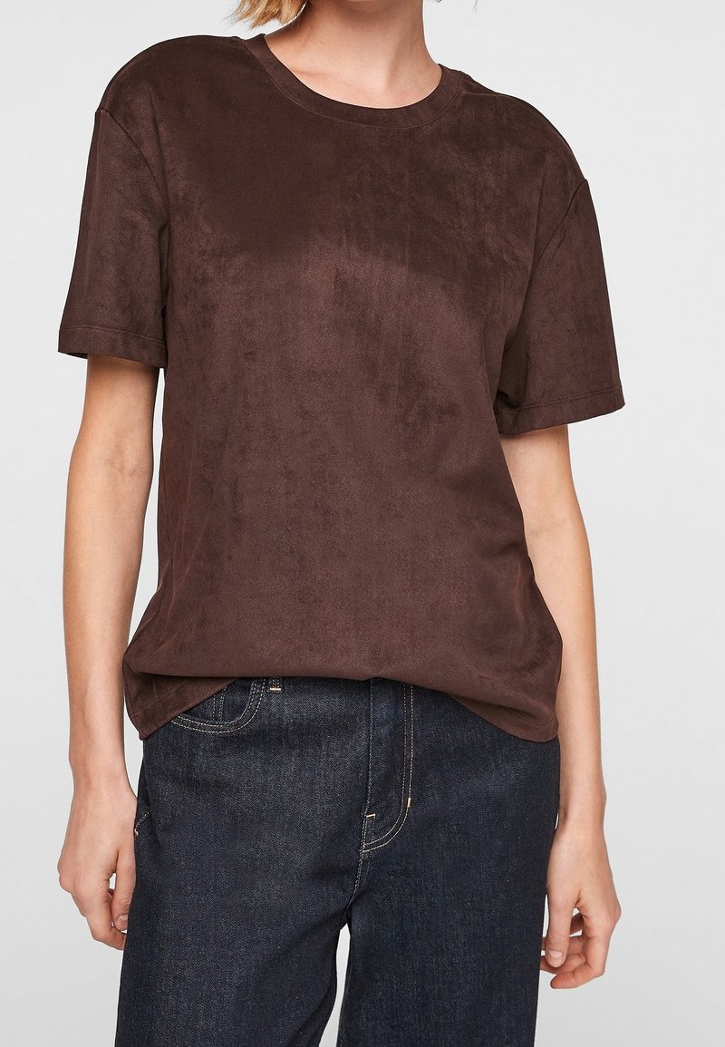 Personne portant un t-shirt marron uni à manches courtes et un jean bleu foncé, les mains détendues le long du corps, sur un fond clair uni.