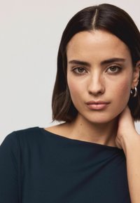 Dark navy top with a round neckline and long sleeves, smooth texture. Model has short dark hair and minimalist hoop earrings.