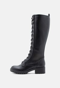 Botte haute en cuir noir jusqu'au genou avec laçage frontal, accessoires en argent, bout arrondi et talon bloc. Fini texturé avec semelle en caoutchouc durable.