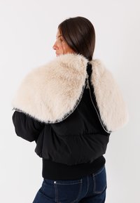 Una donna con lunghi capelli scuri indossa una mantella in pelliccia sintetica beige sopra una giacca nera e jeans blu, rivolta di lato contro uno sfondo bianco.