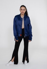 Marineblaue satinierte Bomberjacke, mit Reißverschluss, gerippten Bündchen und Saum. Über einem beigen Oberteil getragen und kombiniert mit schwarzen, ausladenden Hosen mit seitlichen Schlitzen. Weiße Sneaker.