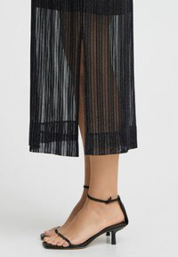 Rochie midi plisată neagră cu textură transparentă și o fente laterală, asortată cu sandale negre cu toc subțire și brâu în jurul gleznei.