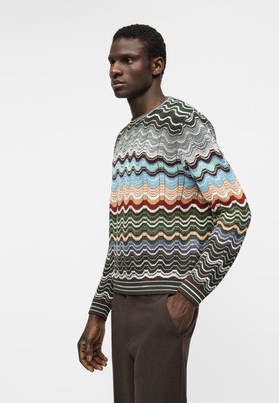 Άνδρας που φοράει πολύχρωμο πουλόβερ με ζιγκ-ζαγκ σχέδιο και καφέ παντελόνι, στέκεται σε γωνιακή στάση τριών τετάρτων μπροστά σε απλό φόντο.