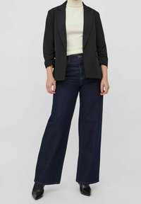 Černý blazer, krémový rolák, široké tmavě modré džíny a černé boty s špičatou špičkou. Blazer má ohrnuté rukávy a přiléhavý střih.