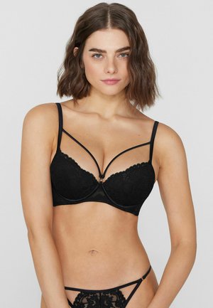 Frau mit schulterlangem braunem Haar, die einen schwarzen Spitzen-BH mit dekorativen Trägern und passende Spitzen-Unterwäsche vor einem schlichten Hintergrund trägt.