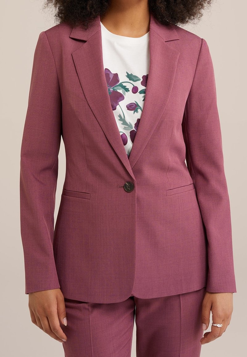 Femme portant un costume mauve ajusté avec un seul bouton, associé à une chemise blanche ornée d'imprimés floraux violets.