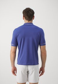 Kortärmad polo i solid lila tyg med en slät textur och ribbad krage; har varumärkeslogotyp på ärmarna.